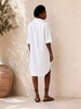 Person wearing a white shirt dress standing against a beige wall with a plant to the left.