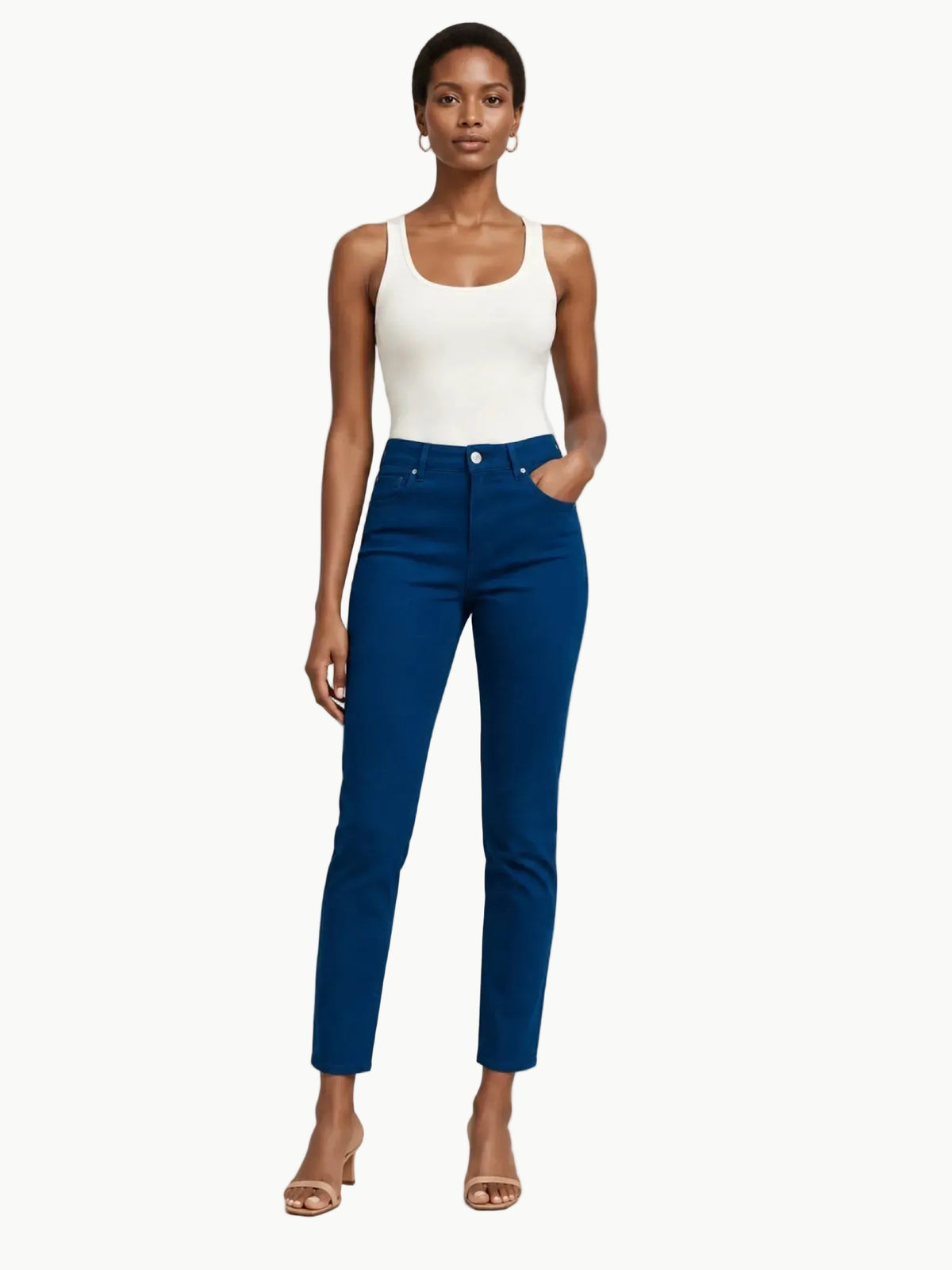 A model stands against a light gray studio background wearing a white sleeveless top and high-waisted, straight-leg blue jeans with her hand in her pocket.