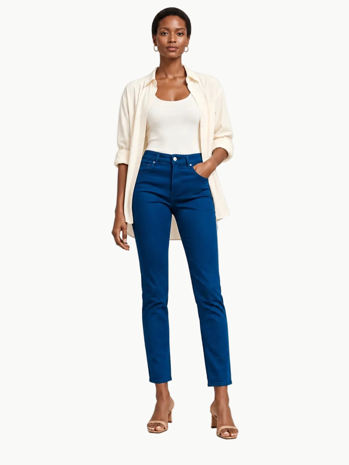A model stands against a light gray studio background wearing a cream button-up shirt over a white tank top, paired with bright blue straight-leg jeans and nude heeled sandals.