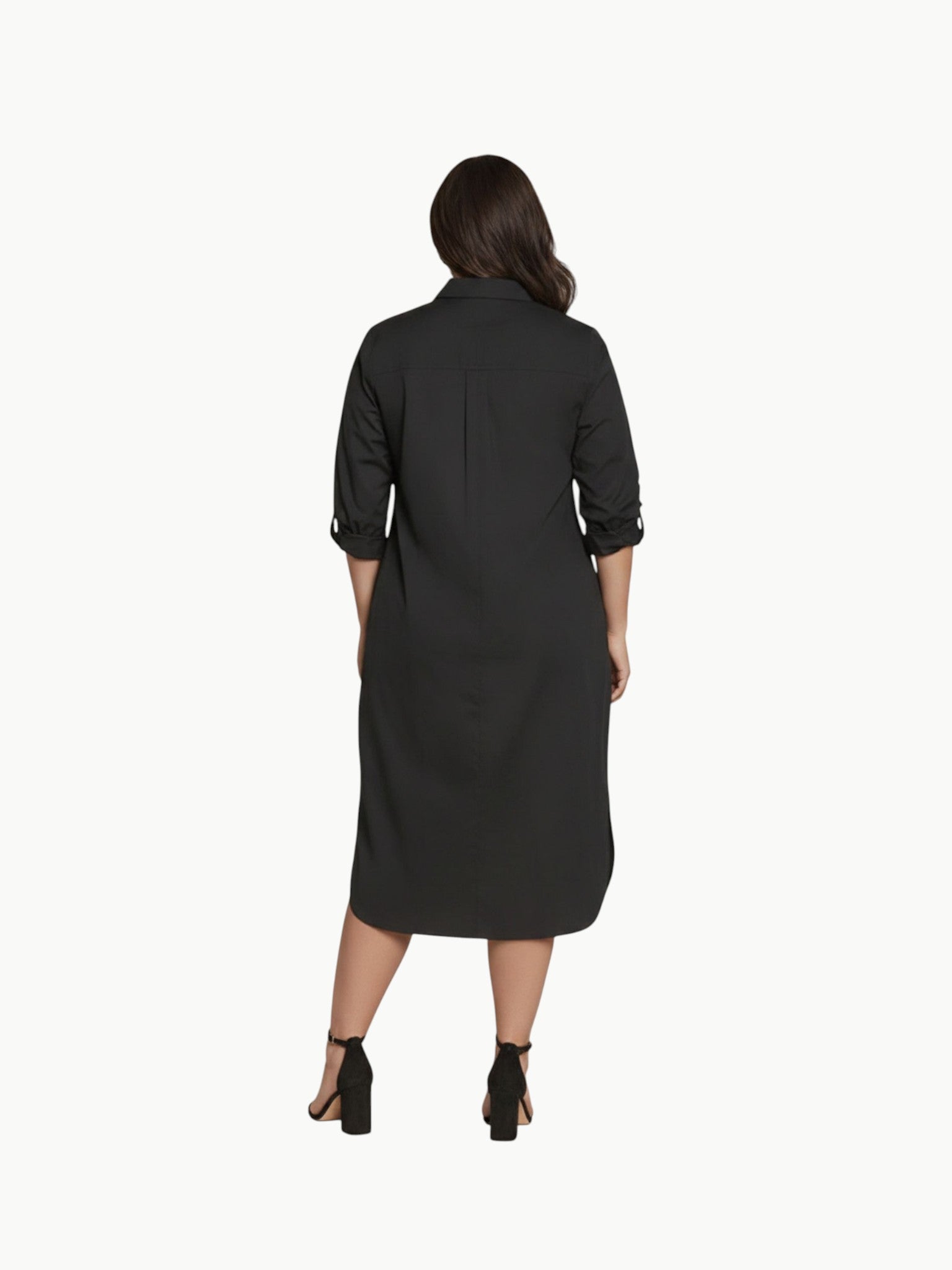 A full-length shot of a plus-size model wearing a black, knee-length stretch poplin shirtdress. The dress features a classic collar, light contrast buttons down the front, a left chest pocket, and side seam pockets where her hands are resting. She is styled with black strappy block heels.