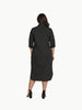 A full-length shot of a plus-size model wearing a black, knee-length stretch poplin shirtdress. The dress features a classic collar, light contrast buttons down the front, a left chest pocket, and side seam pockets where her hands are resting. She is styled with black strappy block heels.