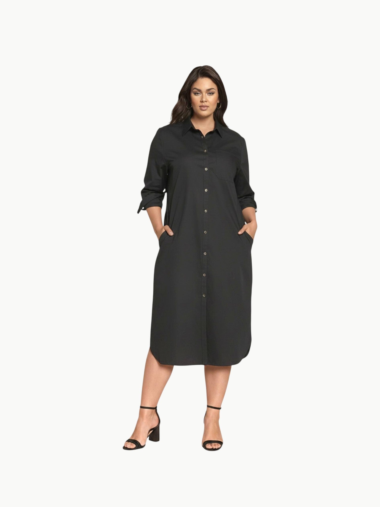 A full-length shot of a plus-size model wearing a black, knee-length stretch poplin shirtdress. The dress features a classic collar, light contrast buttons down the front, a left chest pocket, and side seam pockets where her hands are resting. She is styled with black strappy block heels.