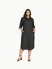 A full-length shot of a plus-size model wearing a black, knee-length stretch poplin shirtdress. The dress features a classic collar, light contrast buttons down the front, a left chest pocket, and side seam pockets where her hands are resting. She is styled with black strappy block heels.