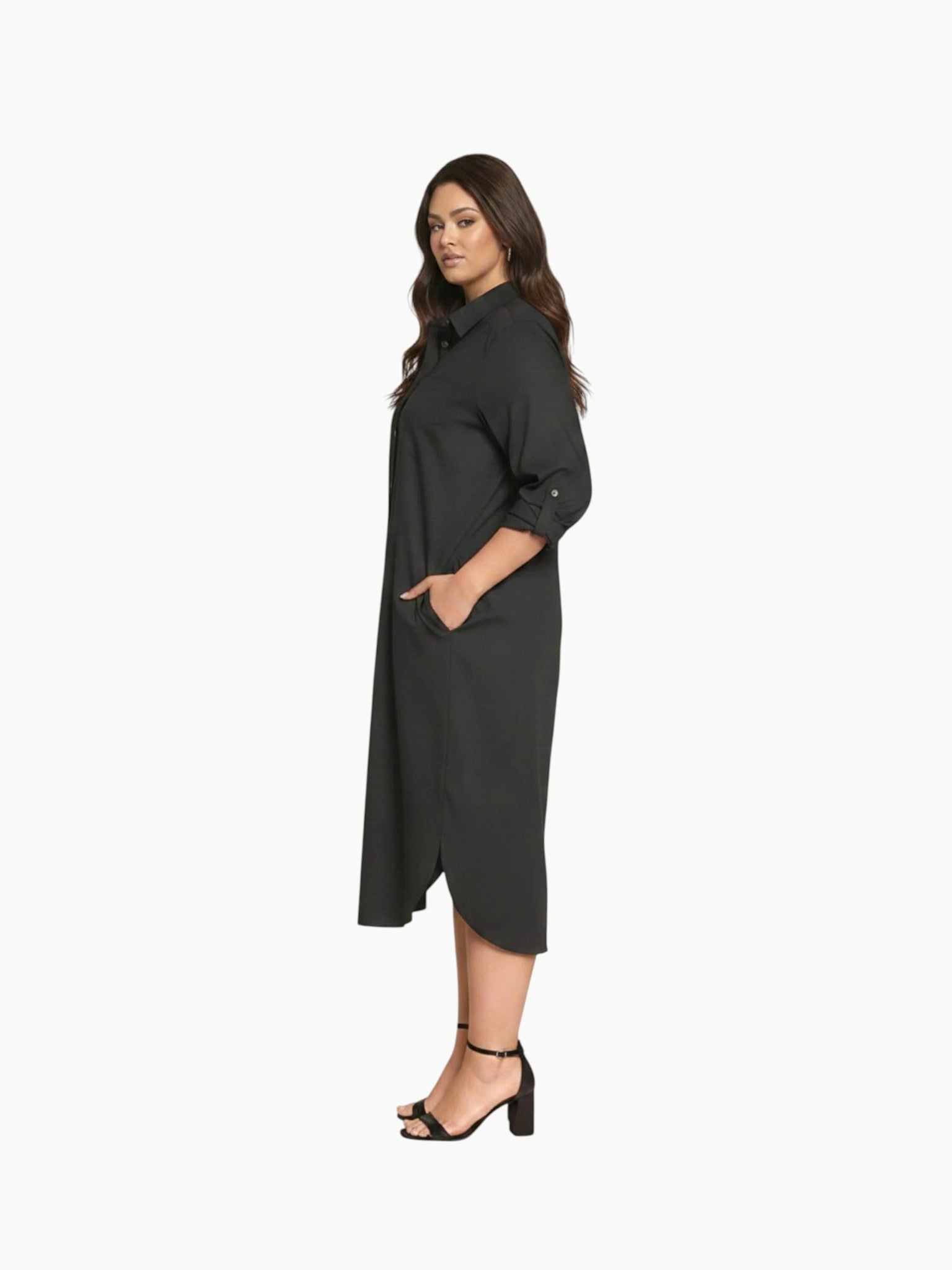 A full-length shot of a plus-size model wearing a black, knee-length stretch poplin shirtdress. The dress features a classic collar, light contrast buttons down the front, a left chest pocket, and side seam pockets where her hands are resting. She is styled with black strappy block heels.