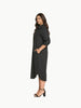 A full-length shot of a plus-size model wearing a black, knee-length stretch poplin shirtdress. The dress features a classic collar, light contrast buttons down the front, a left chest pocket, and side seam pockets where her hands are resting. She is styled with black strappy block heels.