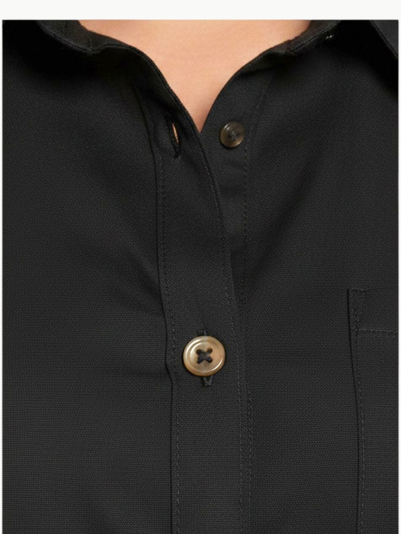 A full-length shot of a plus-size model wearing a black, knee-length stretch poplin shirtdress. The dress features a classic collar, light contrast buttons down the front, a left chest pocket, and side seam pockets where her hands are resting. She is styled with black strappy block heels.