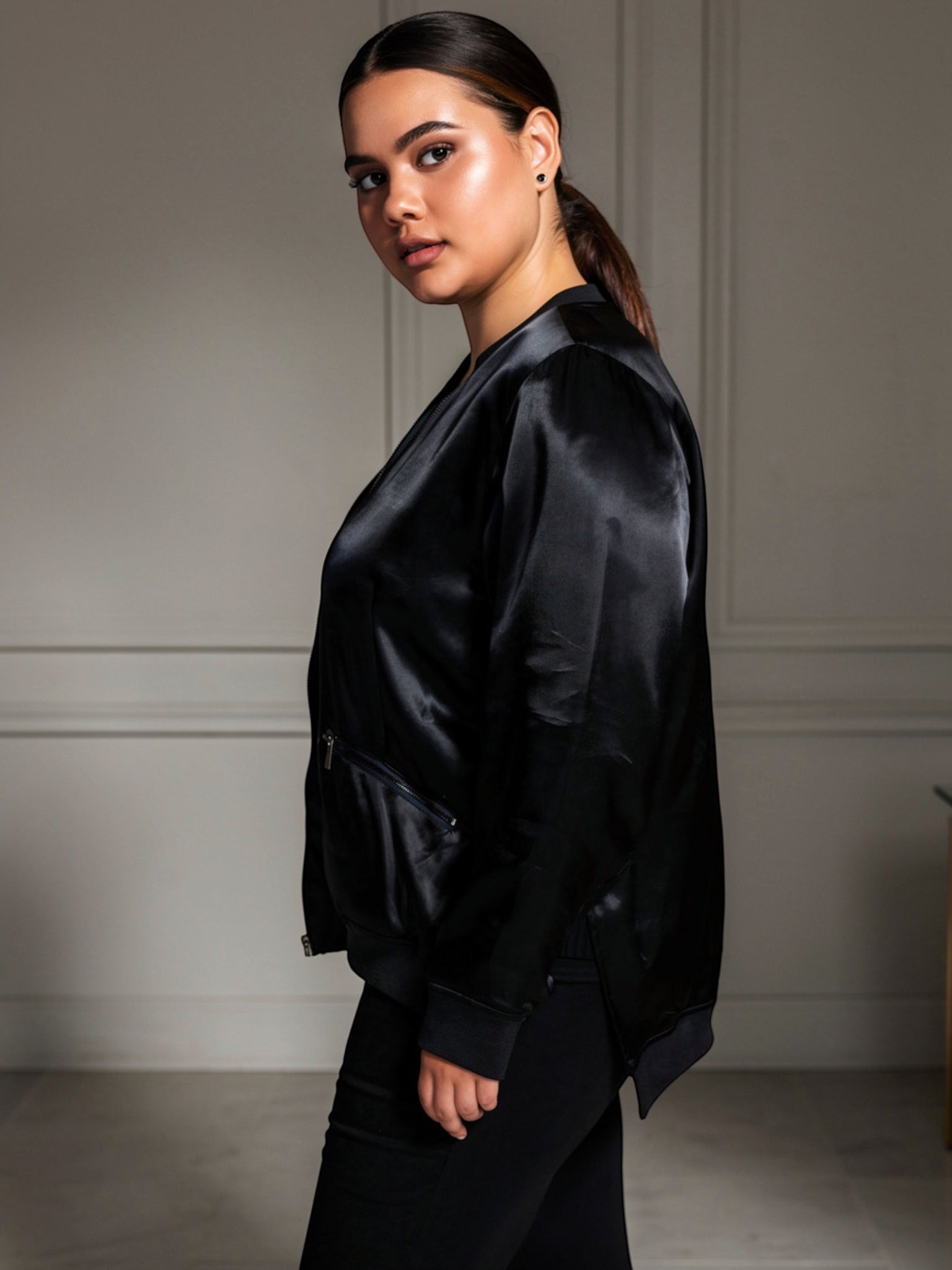 Model wearing a black Queen Anna House of Fashion satin bomber jacket with a high-low hem and zip pockets.